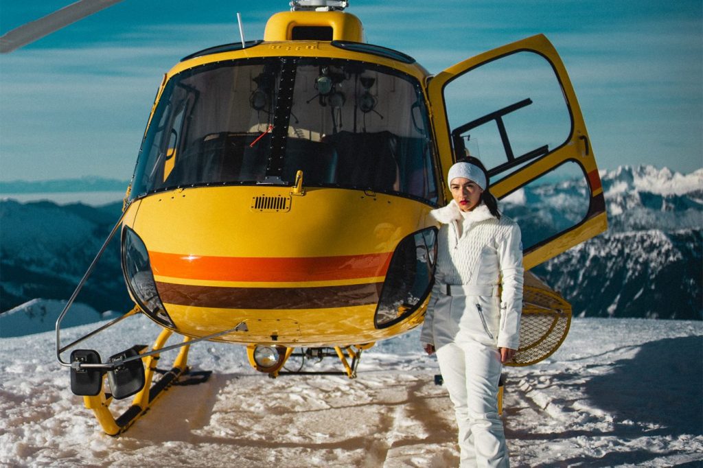 How to Get the Snow Bunny Look for Your Ski Trip. Gabriella wears a white ski suit and stands next to a yellow helicopters on top of a glacier on the west coast of Canada.